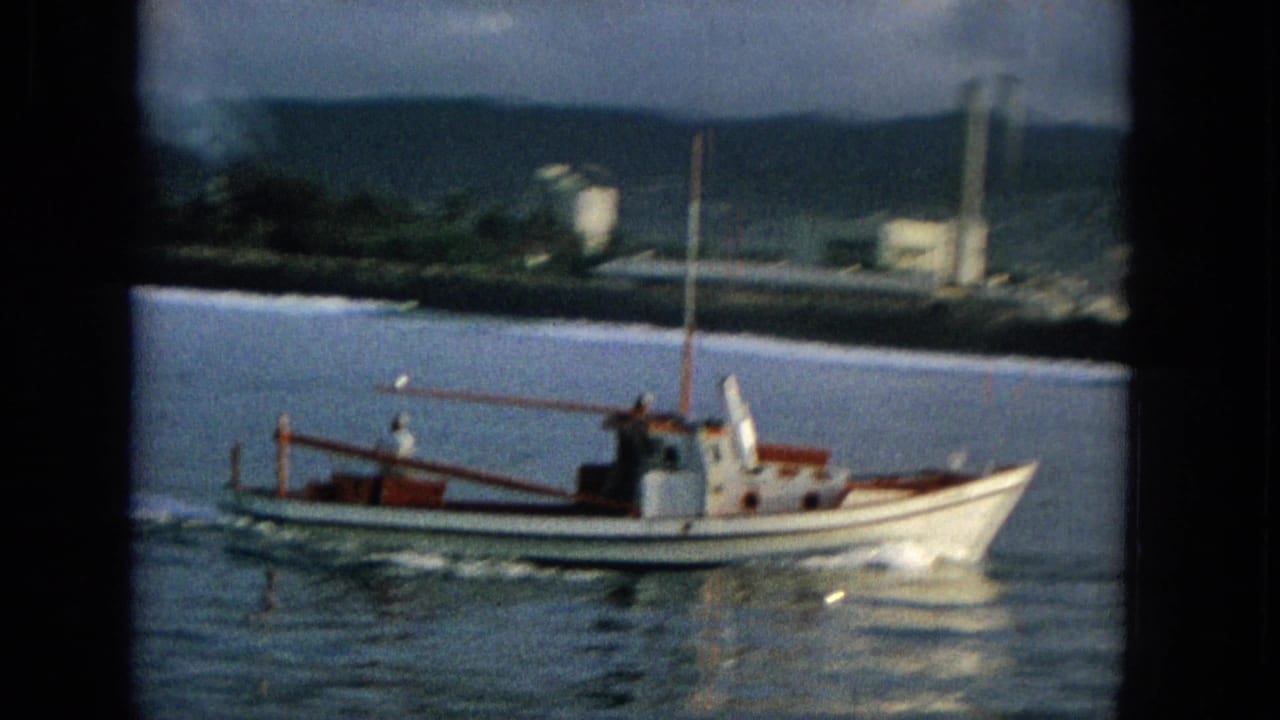 The small white and red boat continues across the water, with a foamy wake and distant shoreline structures set against green hills.