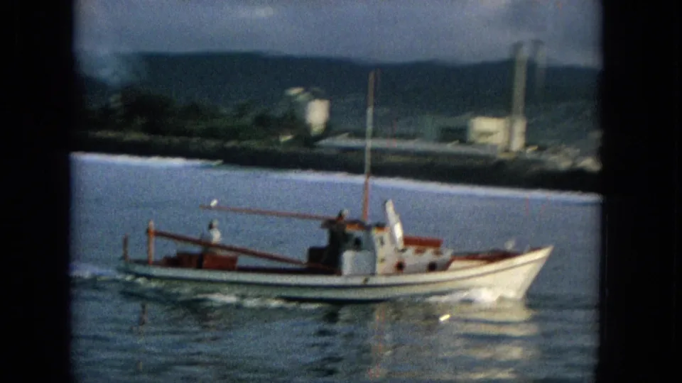 The small white and red boat continues across the water, with a foamy wake and distant shoreline structures set against green hills.