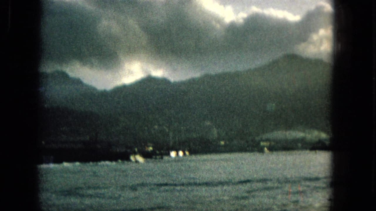 1965 Hawaii 8mm frame showing a dark mountain shoreline across rippling water, with heavy clouds and small lights along the coast.