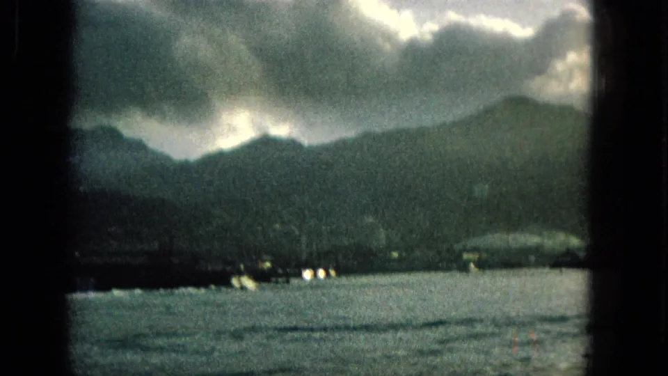 1965 Hawaii 8mm frame showing a dark mountain shoreline across rippling water, with heavy clouds and small lights along the coast.