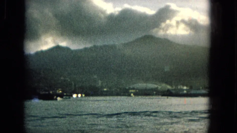1965 Hawaii home-movie frame with distant mountains, a low shoreline, scattered lights, and overcast sky viewed across water from a boat.