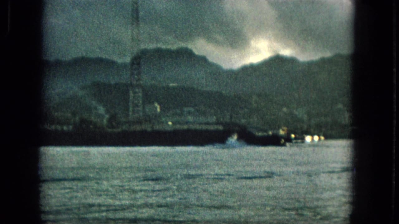 1965 Hawaii 8mm frame showing a waterside strip with a tall tower, distant mountains, shoreline lights, and choppy water beneath dark clouds.