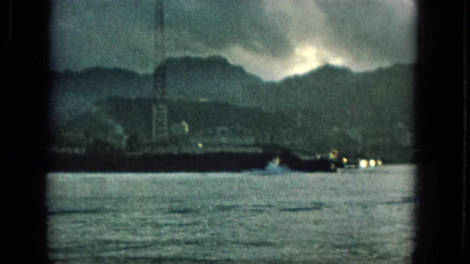 1965 Hawaii 8mm frame showing a waterside strip with a tall tower, distant mountains, shoreline lights, and choppy water beneath dark clouds.