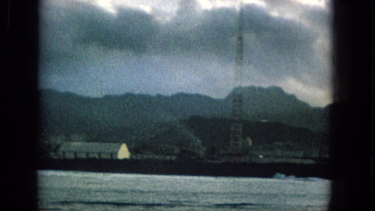 1965 Hawaii home-movie frame with bright water in the foreground, a dark waterfront strip, a tall tower, and mountain ridges below a cloudy sky.