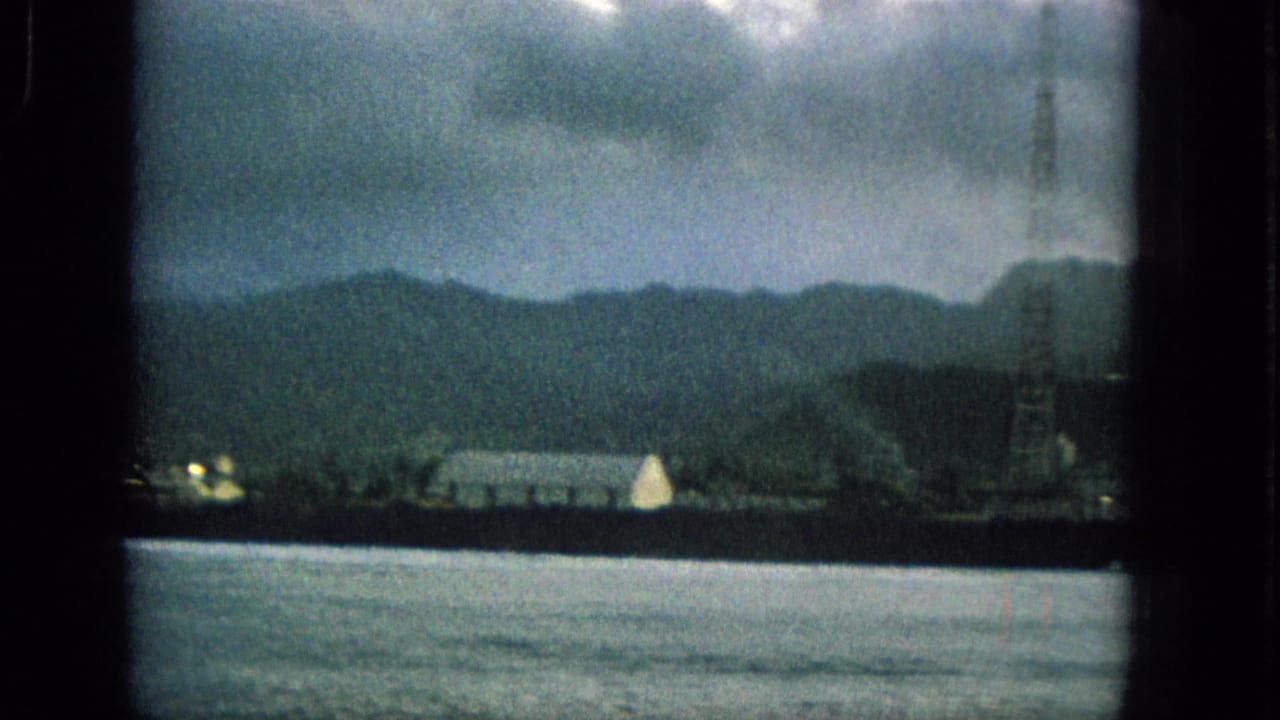 1965 Hawaii 8mm frame showing a cloudy mountainous shoreline, water, distant buildings, and a tower seen through a dark window frame.