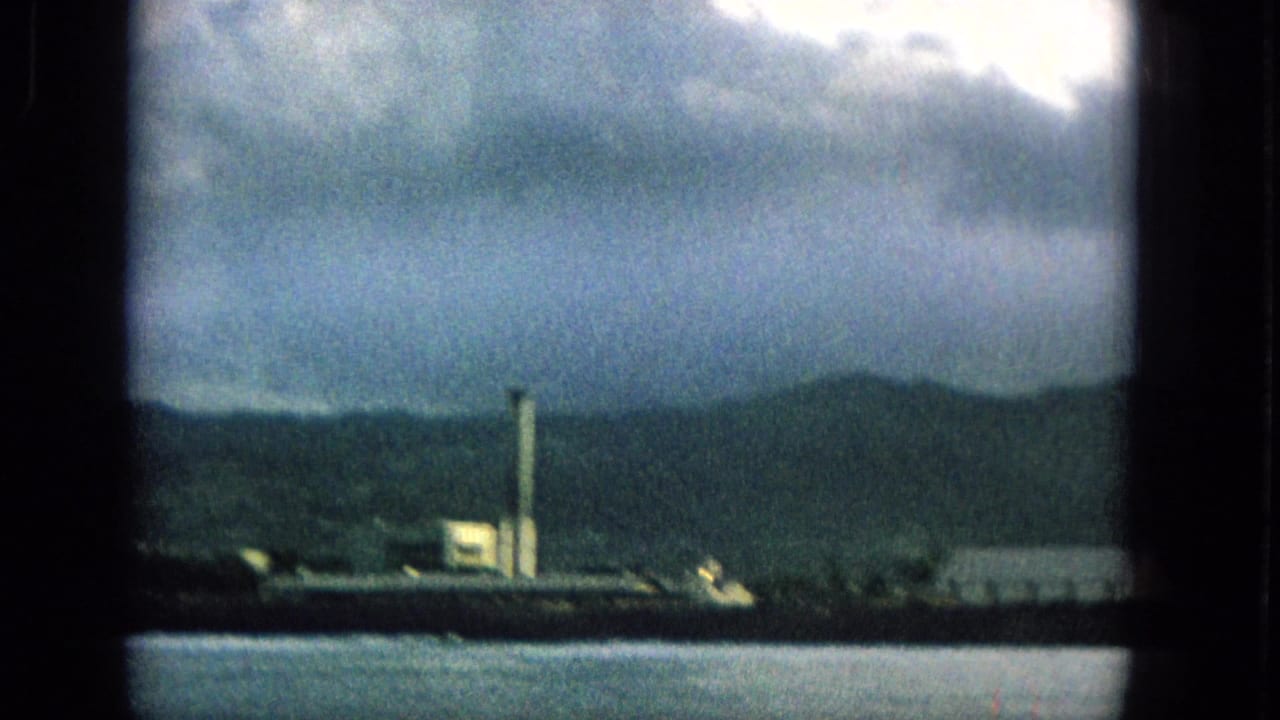 1965 Hawaii 8mm frame of a distant shoreline with mountains, a tall tower-like structure, low buildings, and water viewed through a window.