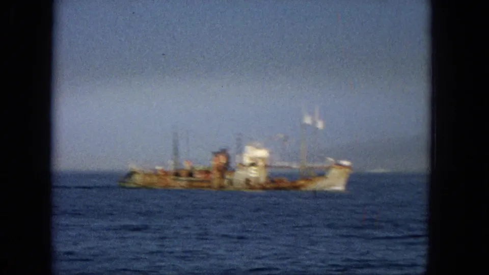 1965 Hawaii 8mm frame showing a rust-streaked ship offshore on blue water with a hazy horizon and pale sky.