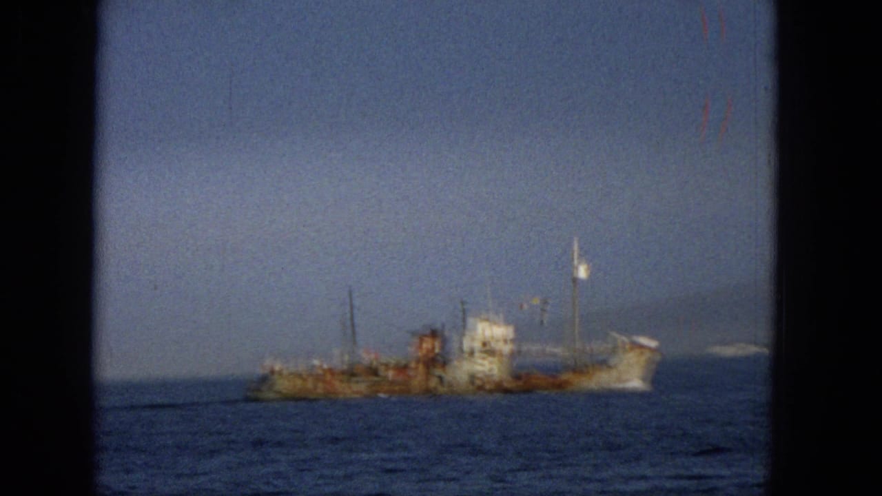 1965 Hawaii 8mm frame of a weathered vessel crossing or floating offshore, with blue water, hazy sky, and a small distant landform visible near the horizon.