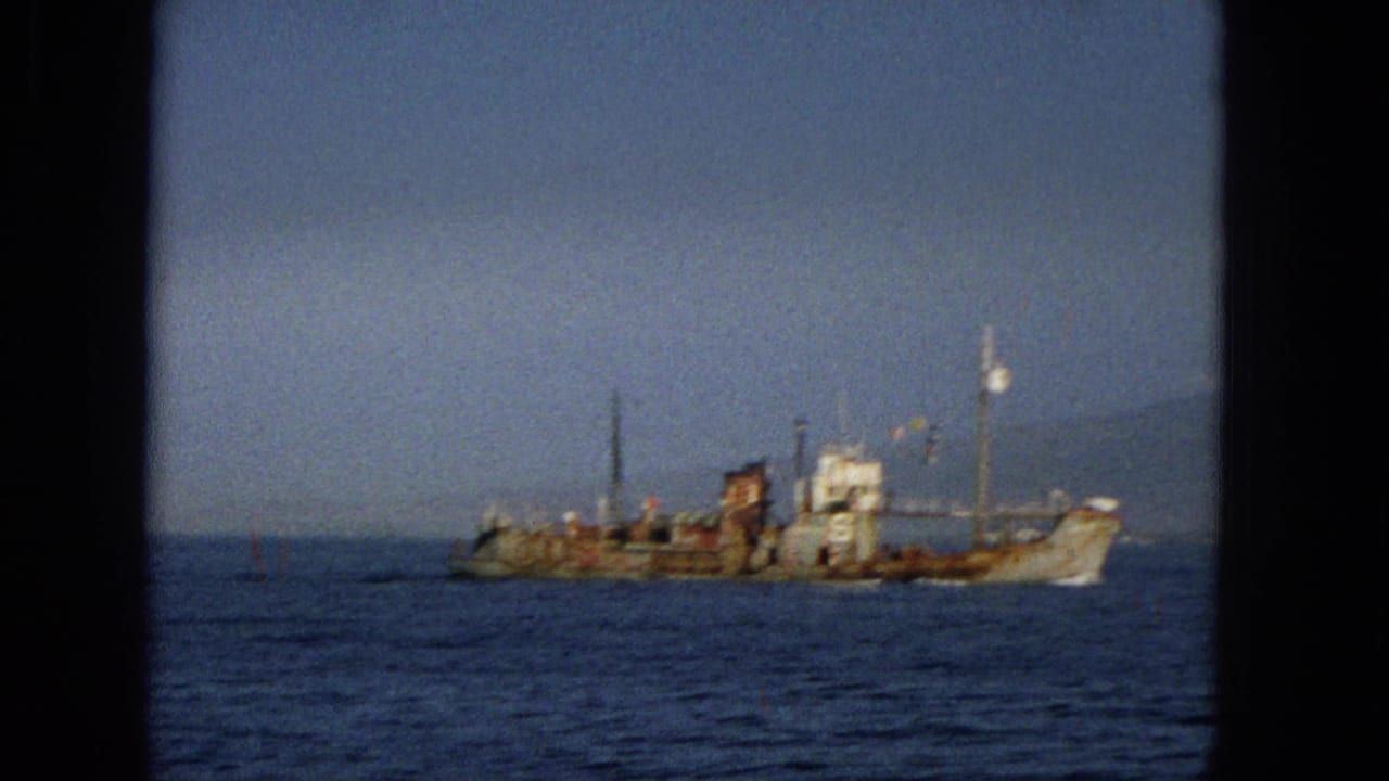 Distant 1965 Hawaii home-movie view of a rusted cargo or utility vessel on the ocean, with a faint shoreline or mountain silhouette behind it.