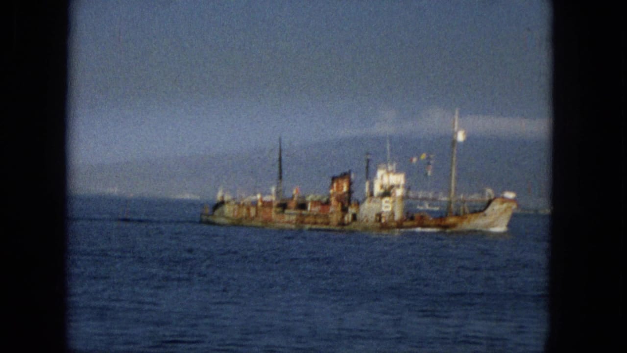 Soft 8mm frame from Hawaii in 1965 showing the side of an aged vessel with visible deck structures, rust staining, and a calm sea foreground.