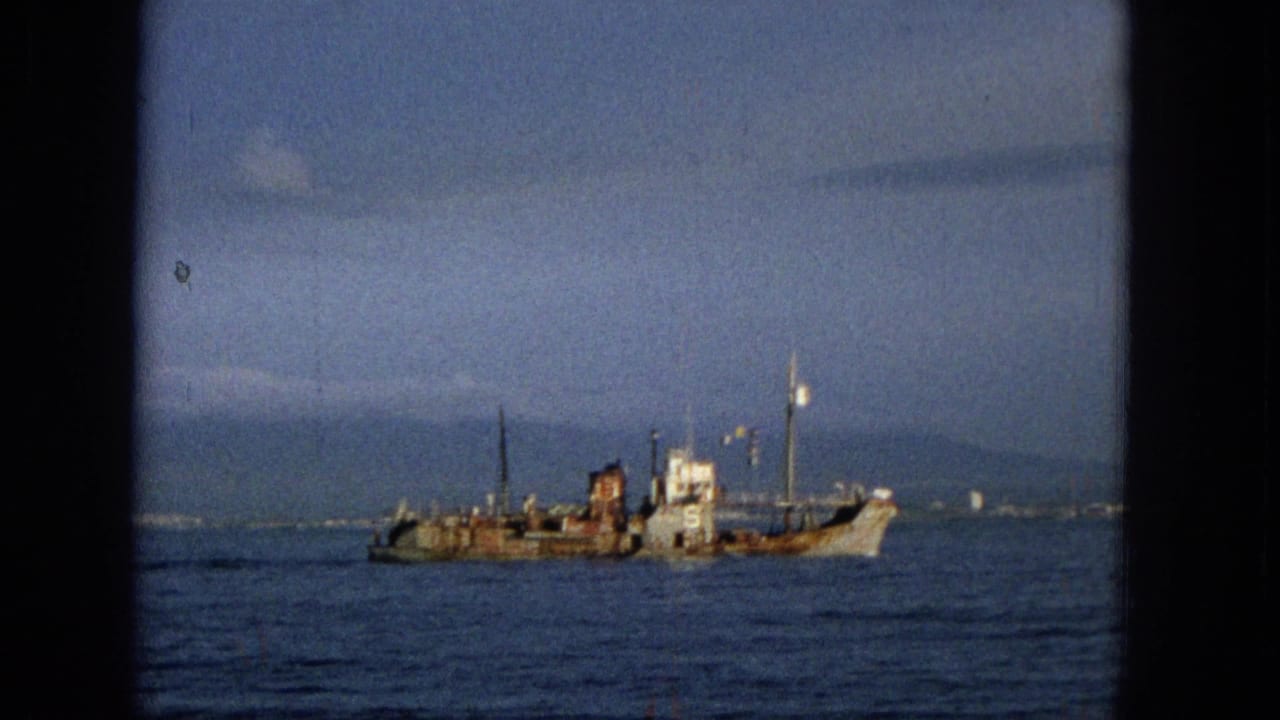 1965 Hawaii 8mm frame showing the same weathered ship moving across blue ocean water, with a faint surf line or shoreline beyond it.