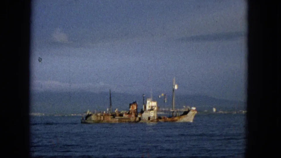 1965 Hawaii 8mm frame showing the same weathered ship moving across blue ocean water, with a faint surf line or shoreline beyond it.