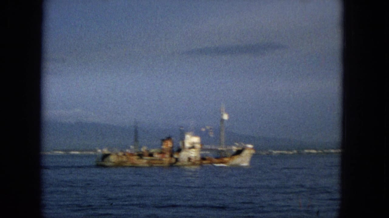 1965 Hawaii home-movie frame with a soft-focus view of a weathered small ship on calm ocean water, framed by dark film edges and a hazy sky.