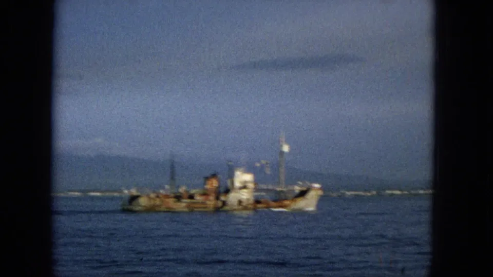 1965 Hawaii home-movie frame with a soft-focus view of a weathered small ship on calm ocean water, framed by dark film edges and a hazy sky.