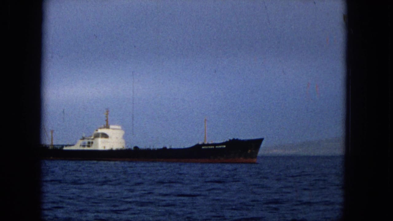 1965 Hawaii 8mm frame showing a large ship with a dark hull and white superstructure moving across calm blue water under a hazy sky.