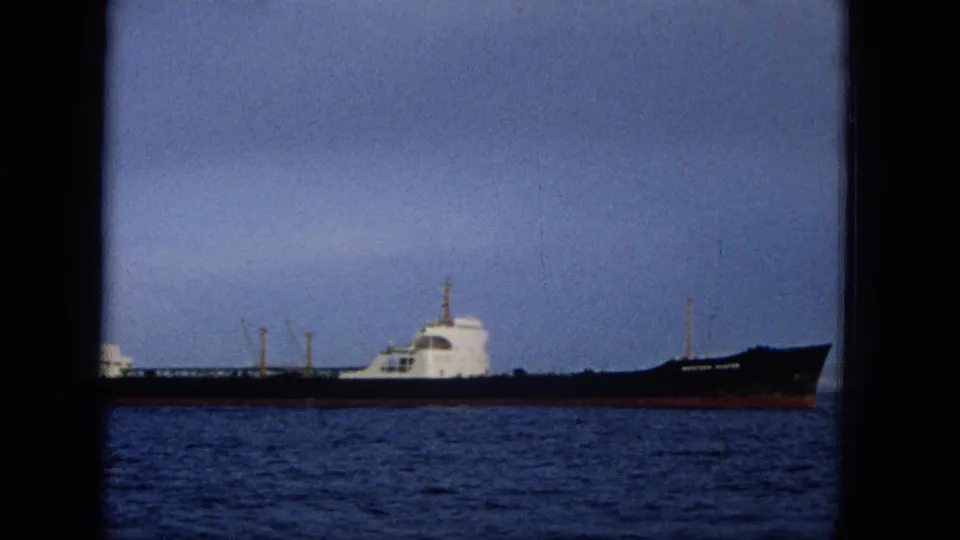 1965 Hawaii 8mm frame with a long dark-hulled ship in profile, white cabin structures, blue water, hazy sky, and visible film-gate edges.