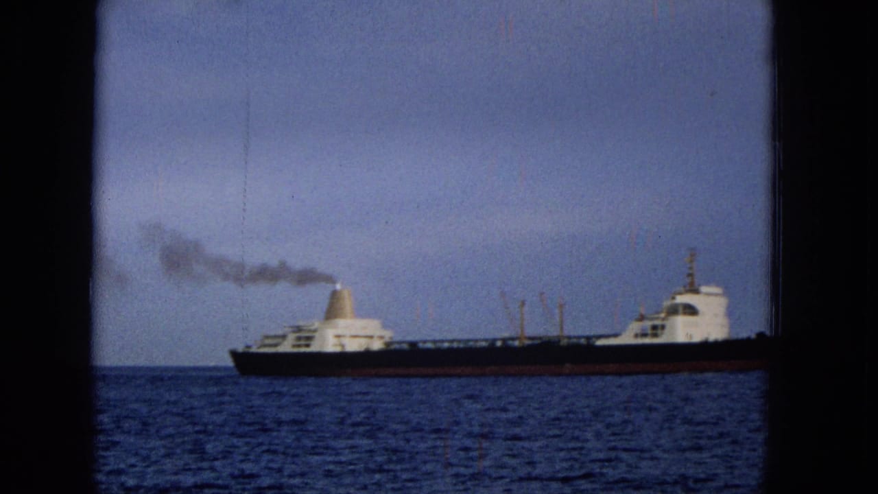 1965 Hawaii 8mm frame showing the vessel farther across the water, with a dark hull, white deck structures, a smoke plume, and a hazy horizon.