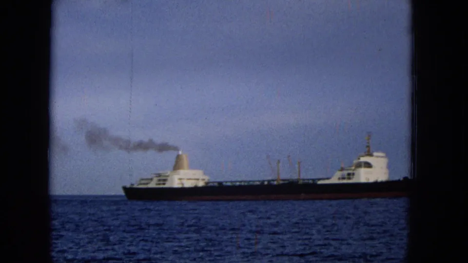 1965 Hawaii 8mm frame showing the vessel farther across the water, with a dark hull, white deck structures, a smoke plume, and a hazy horizon.