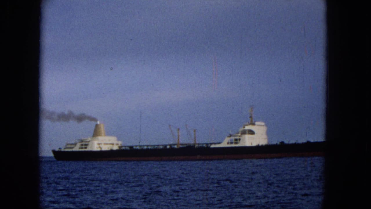 1965 Hawaii home-movie frame showing a long dark-hulled cargo ship with white superstructure moving across blue water under a cloudy sky.