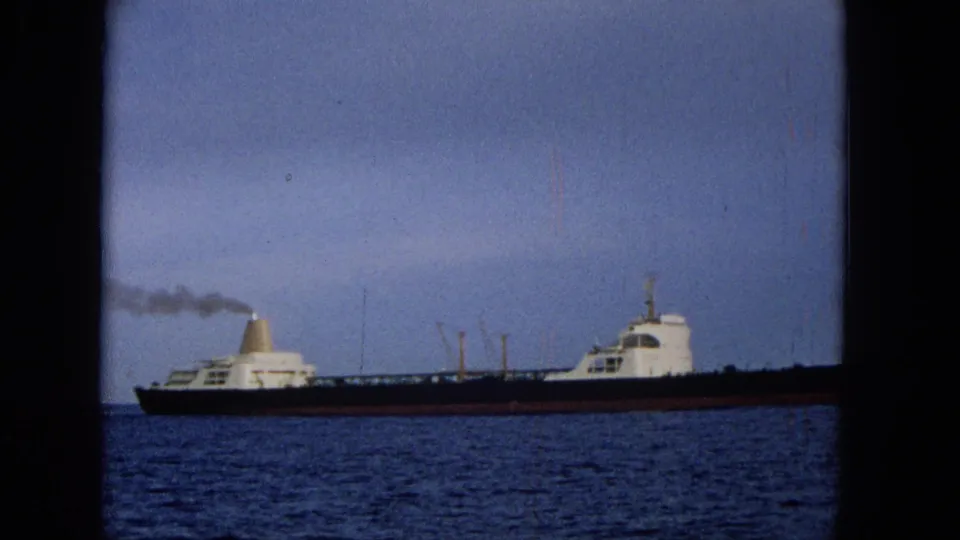 1965 Hawaii home-movie frame showing a long dark-hulled cargo ship with white superstructure moving across blue water under a cloudy sky.