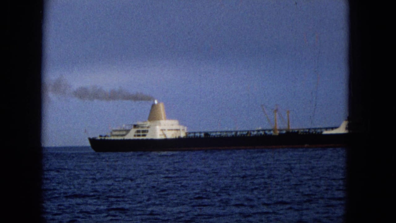 1965 Hawaii home-movie frame with a large cargo vessel in profile, its pale funnel releasing dark smoke over the open ocean.