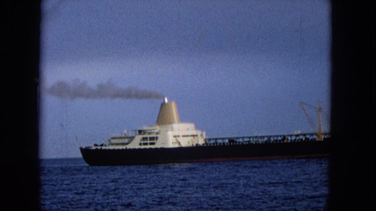 Closer 1965 Hawaii frame of a cargo ship passing at sea, with white upper decks, a pale funnel, and smoke trailing into the sky.
