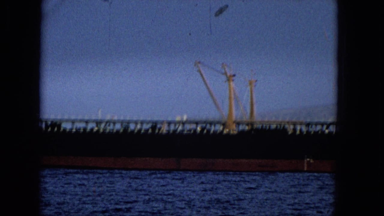 1965 Hawaii 8mm frame showing the tanker's long dark hull and deck railings with distant port cranes visible beyond the vessel.
