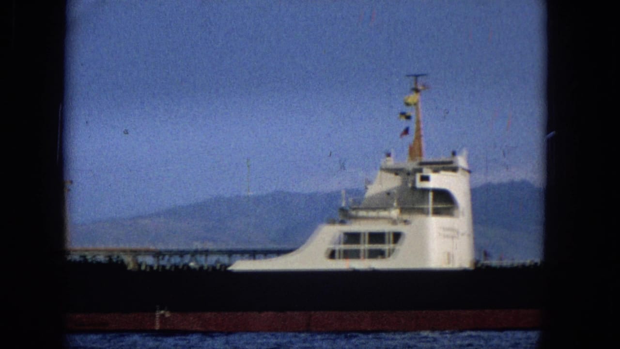1965 Hawaii 8mm frame showing the tanker's white superstructure and signal flags against blue water, sky, and a distant mountain shoreline.