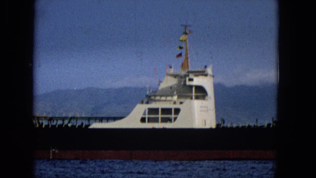 A white ferry or boat moves across blue water with a hazy distant shoreline, seen within the rounded border of 8mm home-movie film.