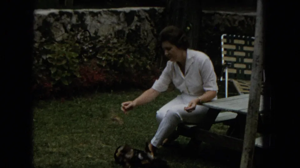 The woman remains seated at the picnic table as the small dog stands or moves close to her feet in the grassy backyard, with a lawn chair and stone wall behind her.