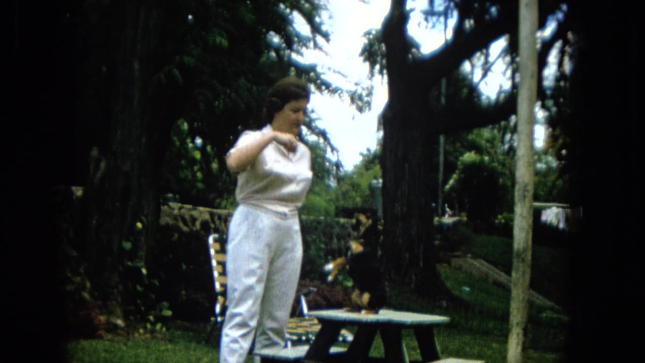 A woman in white stands beside a picnic table as a small black-and-tan dog waits on the tabletop in the 1965 Hawaii garden scene.