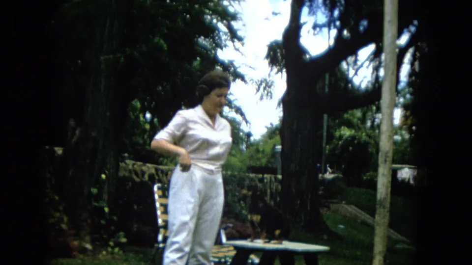 The woman continues the outdoor pet-training session beside the picnic table, with the small dog partly visible against the shaded garden background.