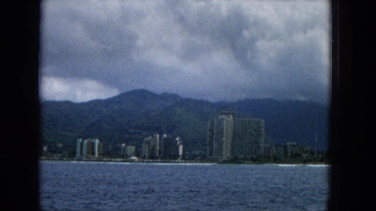 1965 Hawaii home-movie frame showing ocean water in the foreground, a shoreline with tall buildings, green mountains, and dense clouds overhead.