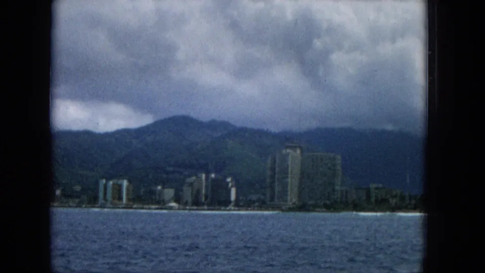 1965 Hawaii home-movie frame showing ocean water in the foreground, a shoreline with tall buildings, green mountains, and dense clouds overhead.