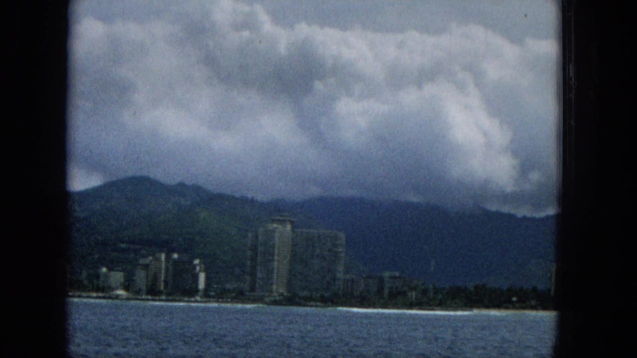 1965 Hawaii coastal view from the water with beachfront buildings, a prominent tower, mountain slopes, and rolling cloud cover.