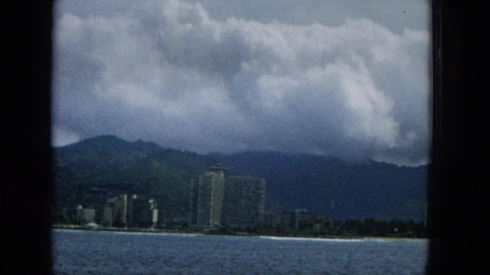 1965 Hawaii coastal view from the water with beachfront buildings, a prominent tower, mountain slopes, and rolling cloud cover.