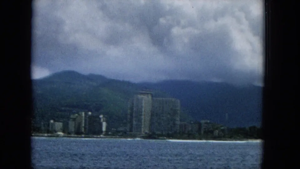 1965 Hawaii frame with blue ocean in front, a strip of beach, several shoreline buildings, and dark mountains under layered clouds.