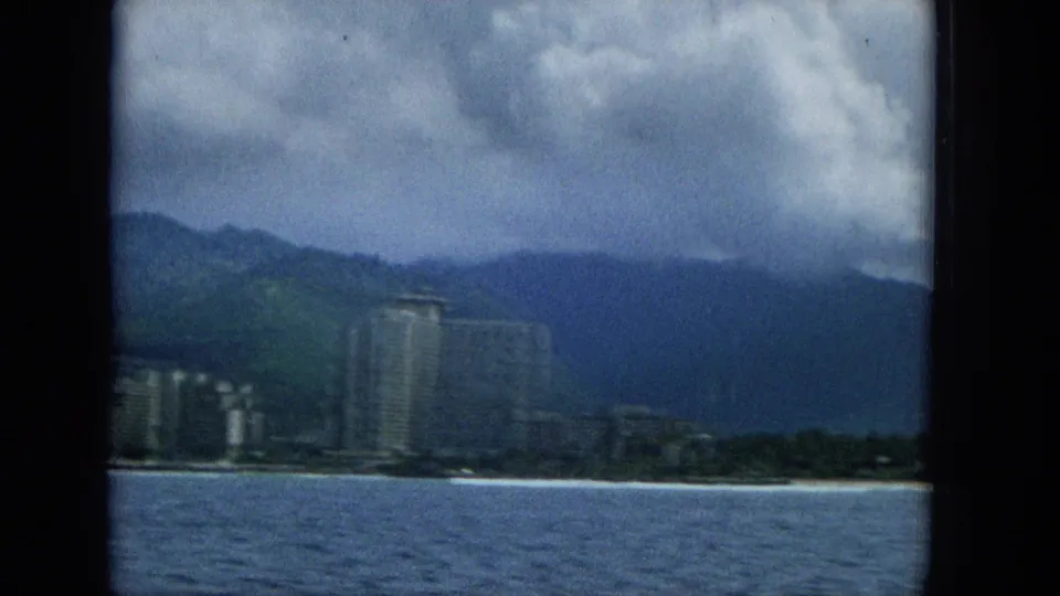1965 Hawaii home-movie frame showing a slightly blurred offshore view of a tall oceanfront building, beach edge, mountain backdrop, and heavy clouds.