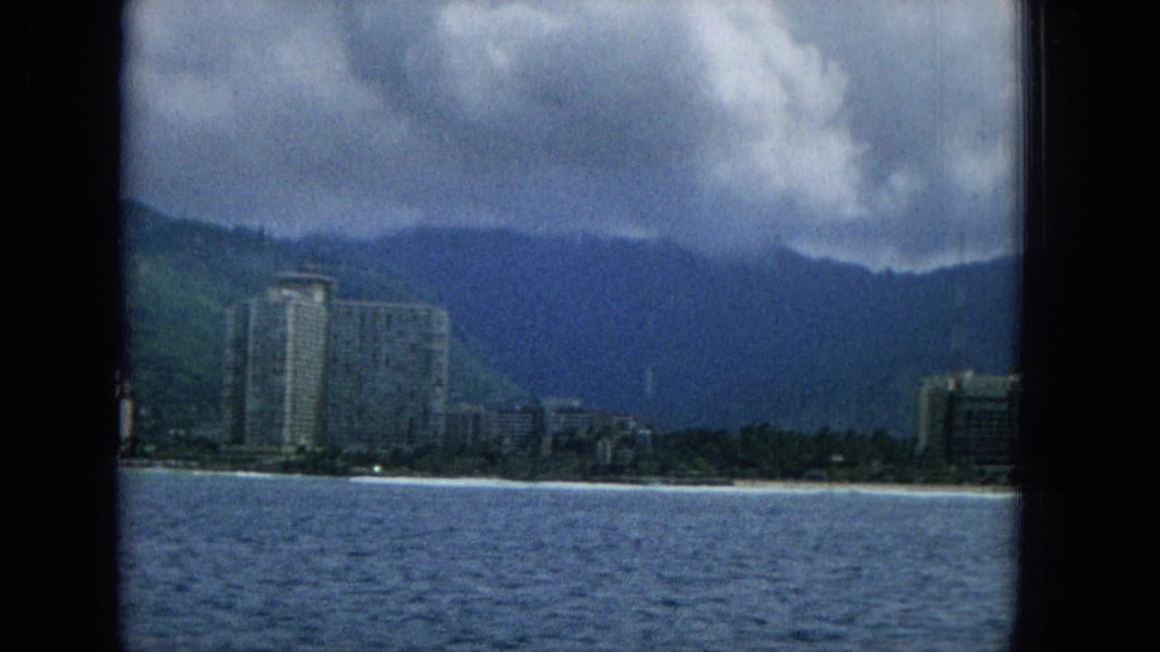 1965 Hawaii 8mm frame looking from the water toward beachfront high-rises, a narrow sandy shore, green mountains, and a cloudy sky.