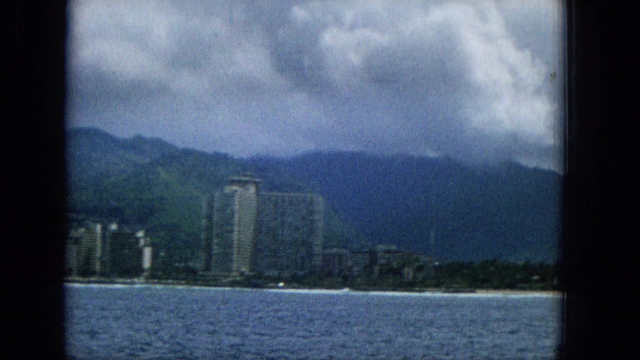 1965 Hawaii travel frame showing resort towers and lower beachfront structures across calm water, backed by green ridges and overcast clouds.