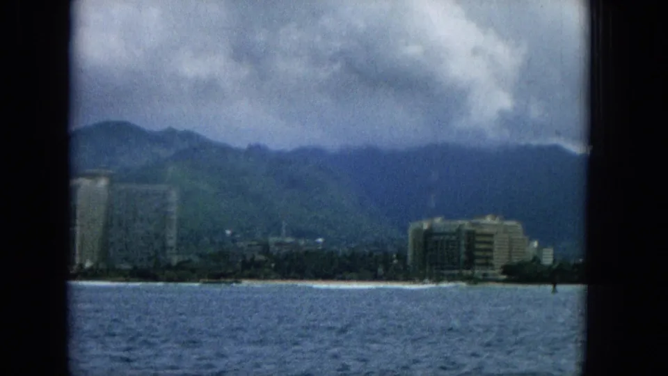 1965 Hawaii 8mm frame with rippled ocean in the foreground, partial high-rise buildings at the beach, and cloud-covered green mountains behind.