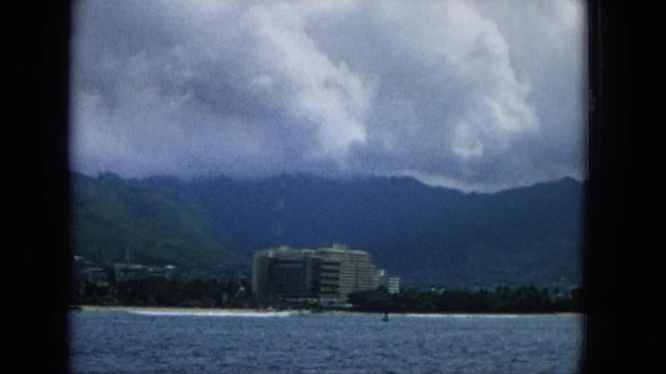 1965 Hawaii vacation-film frame showing a low resort building near the beach, ocean water in front, and dark green mountains beneath heavy clouds.