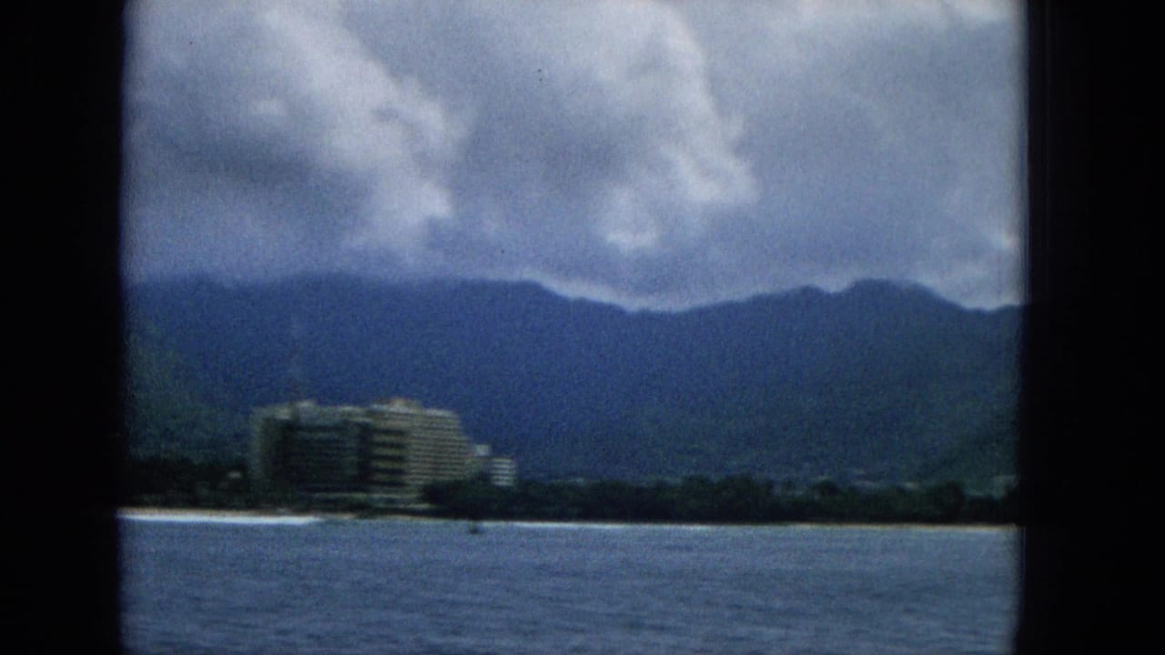 1965 Hawaii 8mm frame from a boat showing open water, a distant resort-like coastal building, palm-lined shore, mountains, and a cloudy sky.
