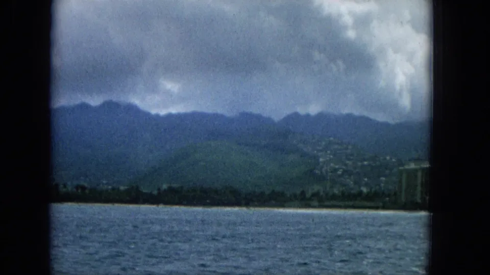 1965 Hawaii 8mm frame showing blue water in the foreground, a narrow beach, green mountains, cloud cover, and distant coastal buildings.