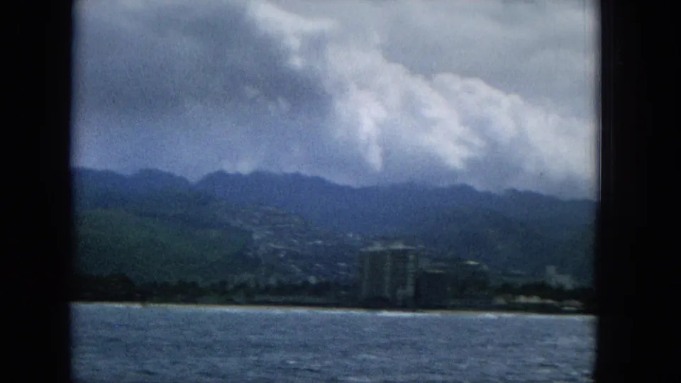 1965 Hawaii 8mm frame from offshore showing rippled water, beachfront buildings, a green mountain backdrop, and a bright cloud bank.