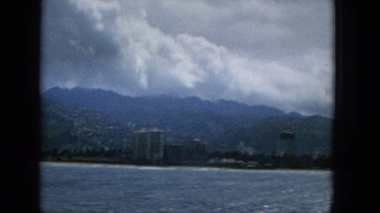 1965 Hawaii 8mm frame showing choppy ocean in the foreground, a low shoreline with a taller resort-style building, and green mountains under heavy clouds.
