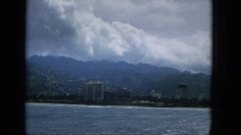 1965 Hawaii 8mm frame showing choppy ocean in the foreground, a low shoreline with a taller resort-style building, and green mountains under heavy clouds.