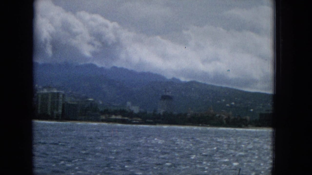 1965 Hawaii shoreline view from a moving boat, with ocean water, pale beach, clustered coastal buildings, mountains, and a dramatic cloud layer.