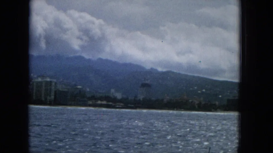 1965 Hawaii shoreline view from a moving boat, with ocean water, pale beach, clustered coastal buildings, mountains, and a dramatic cloud layer.