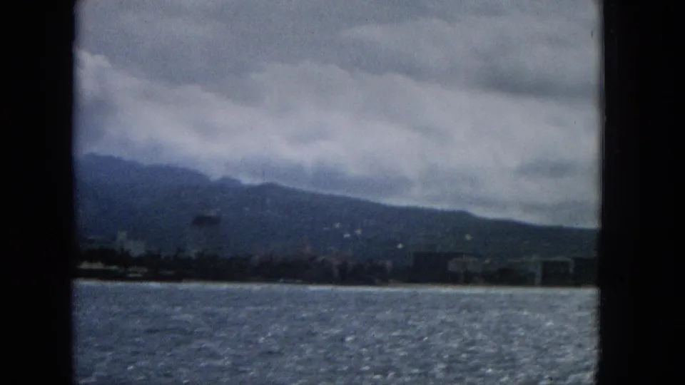 1965 Hawaii 8mm frame with rippled water filling the foreground, a narrow beach and distant buildings along the shore, and mountains partly covered by clouds.